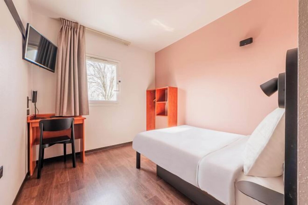 A bedroom with a bed, a desk with a chair, a TV mounted on the wall, and a window with curtains.