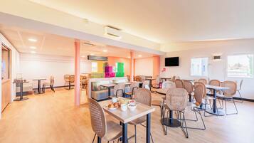 A spacious dining area with multiple tables and chairs, a kitchen area in the background, and a flat-screen TV mounted on the wall.
