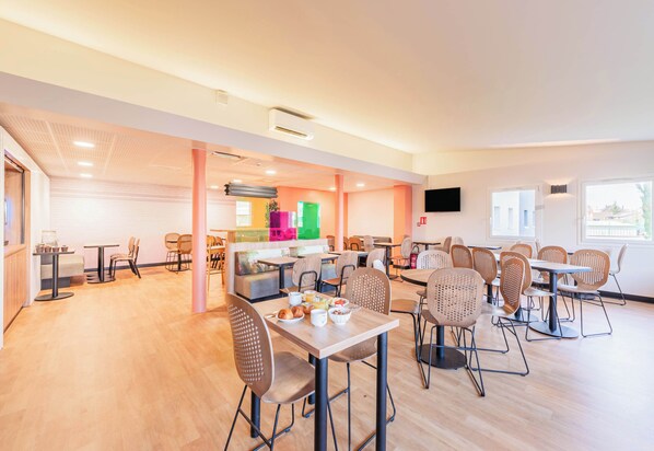 A spacious dining area with multiple tables and chairs, a kitchen area in the background, and a flat-screen TV mounted on the wall.