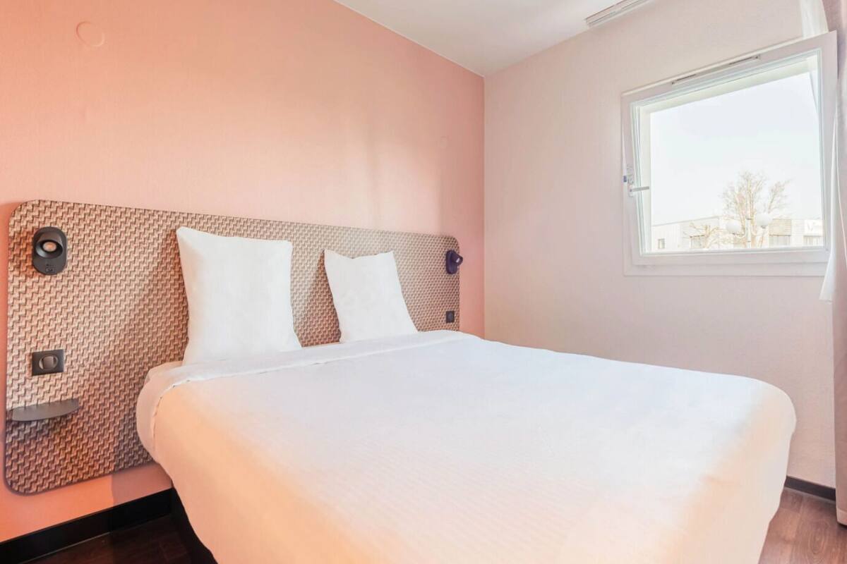Una cama bien hecha con cabecera, una ventana con cortinas y un panel de control instalado en la pared.