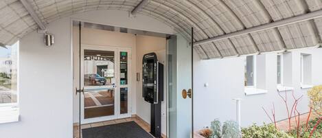 Entrada moderna de un edificio con puerta de cristal, una máquina de café y un pequeño jardín.