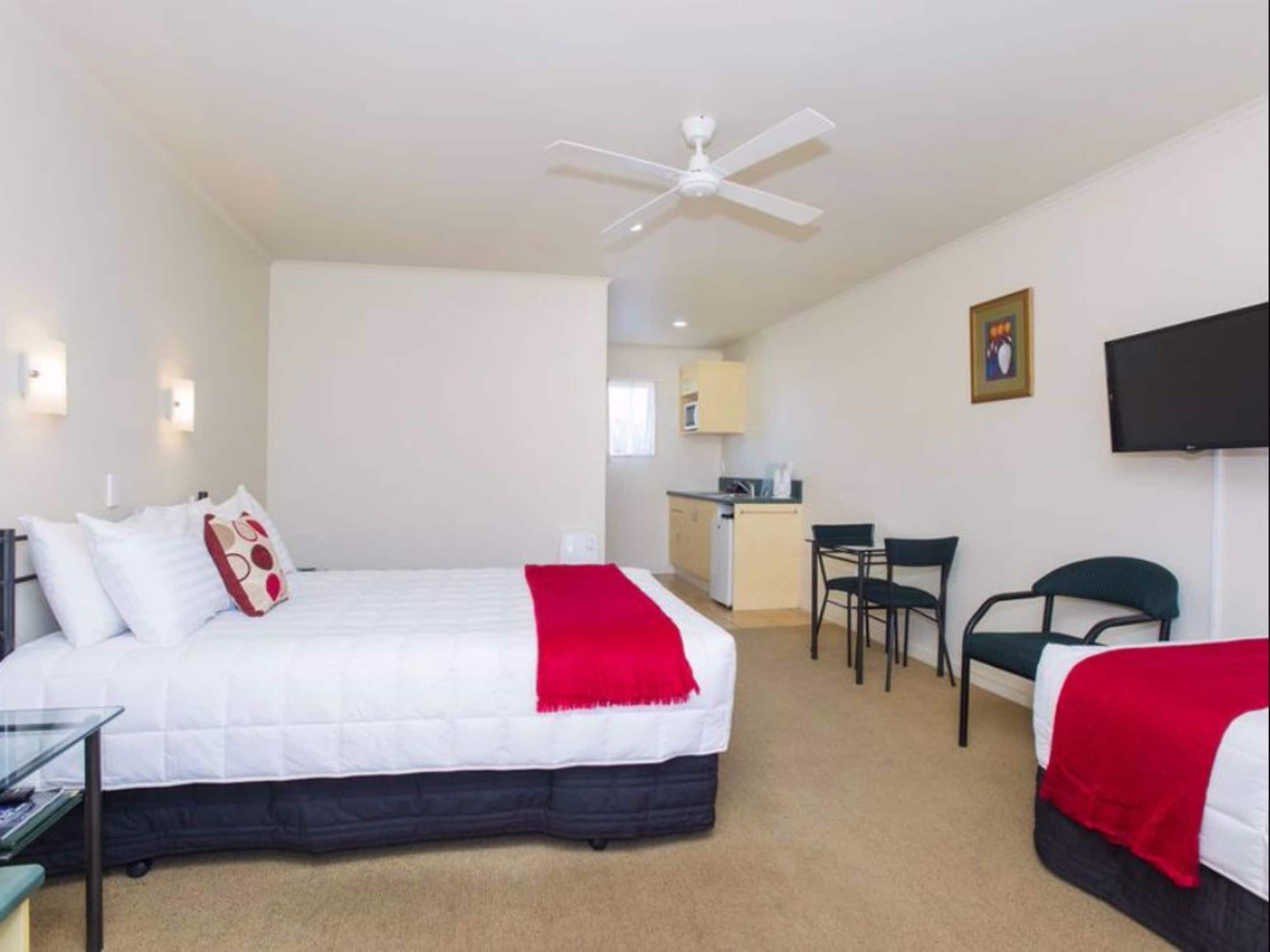 A hotel room with a bed, a desk, a chair, a ceiling fan, a television, and a kitchenette in the background.