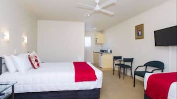 A hotel room with a bed, a desk, a chair, a ceiling fan, a television, and a kitchenette in the background.