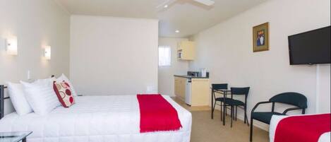 A hotel room with a bed, a desk, a chair, a ceiling fan, a television, and a kitchenette in the background.