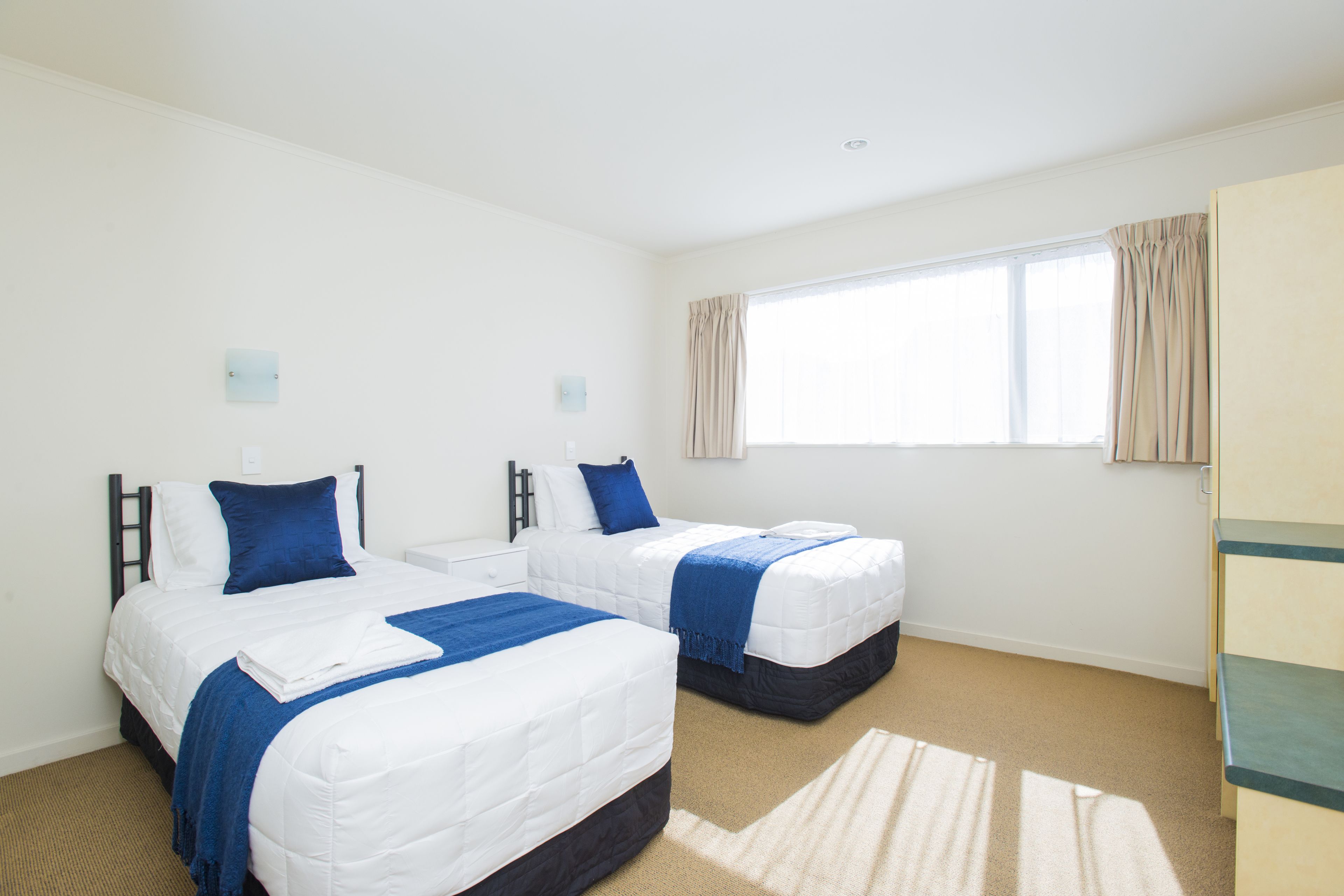 A hotel room with two beds, a large window with curtains, a carpeted floor, and a wardrobe.