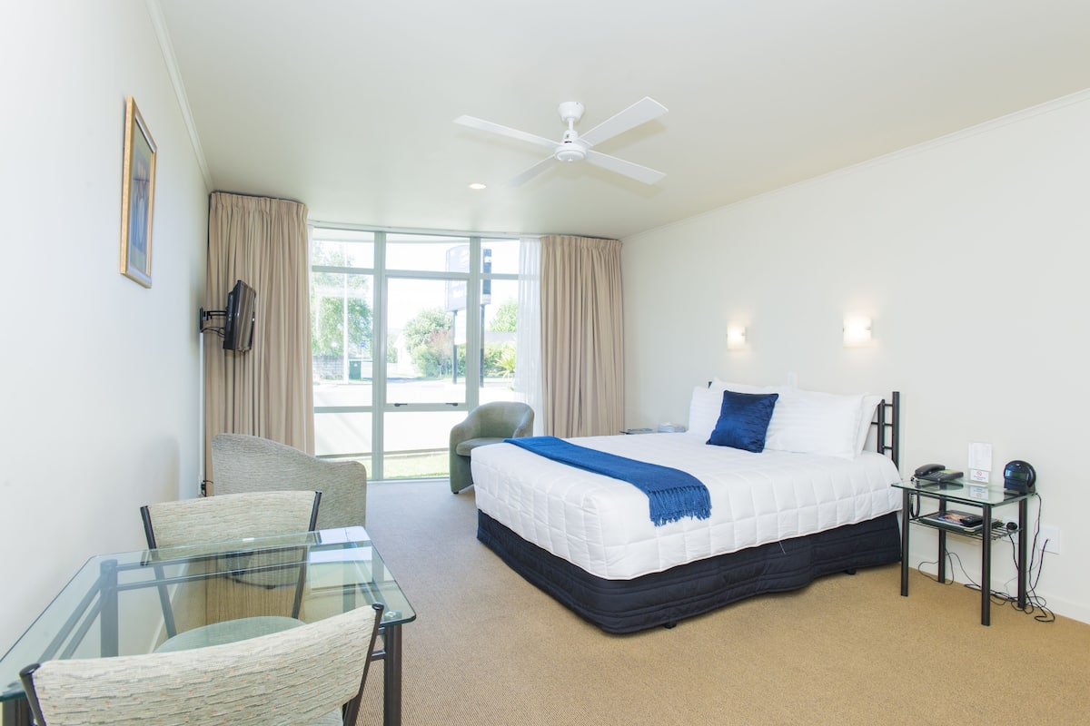 A hotel room with a bed, a glass table, a chair, a desk, a ceiling fan, and a window with curtains.