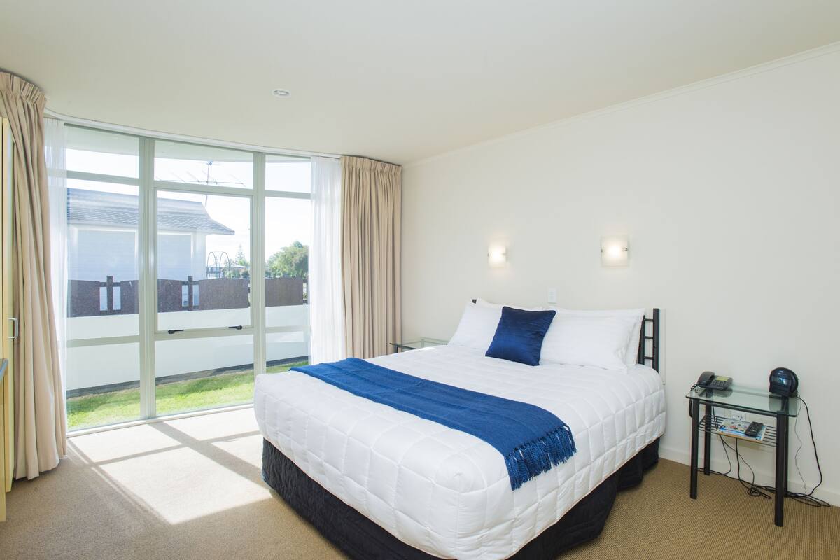 A bedroom with a large bed, a glass door leading to a balcony, and a small table with a phone and a lamp.