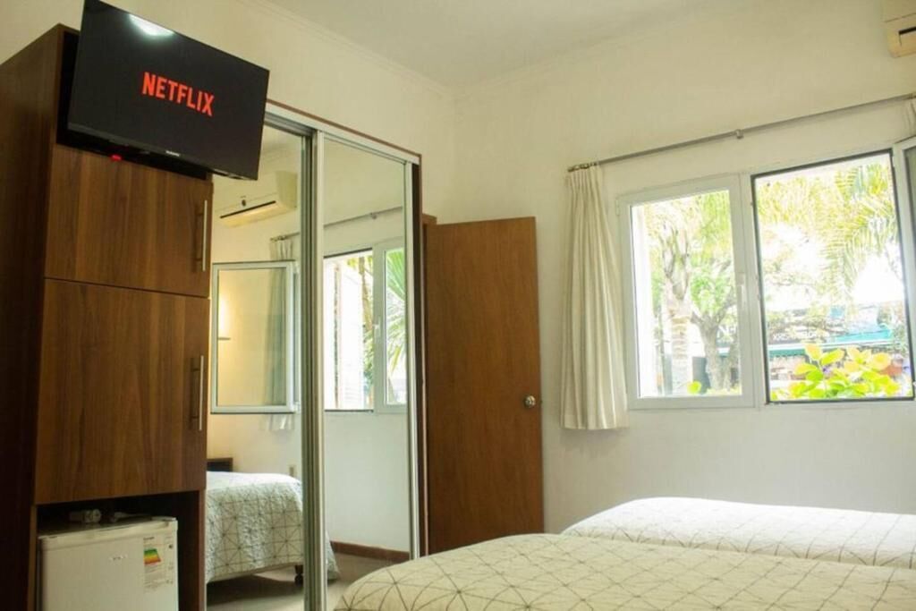 Un dormitorio con cama, armario, televisor sobre un mueble bajo y ventana con cortinas.