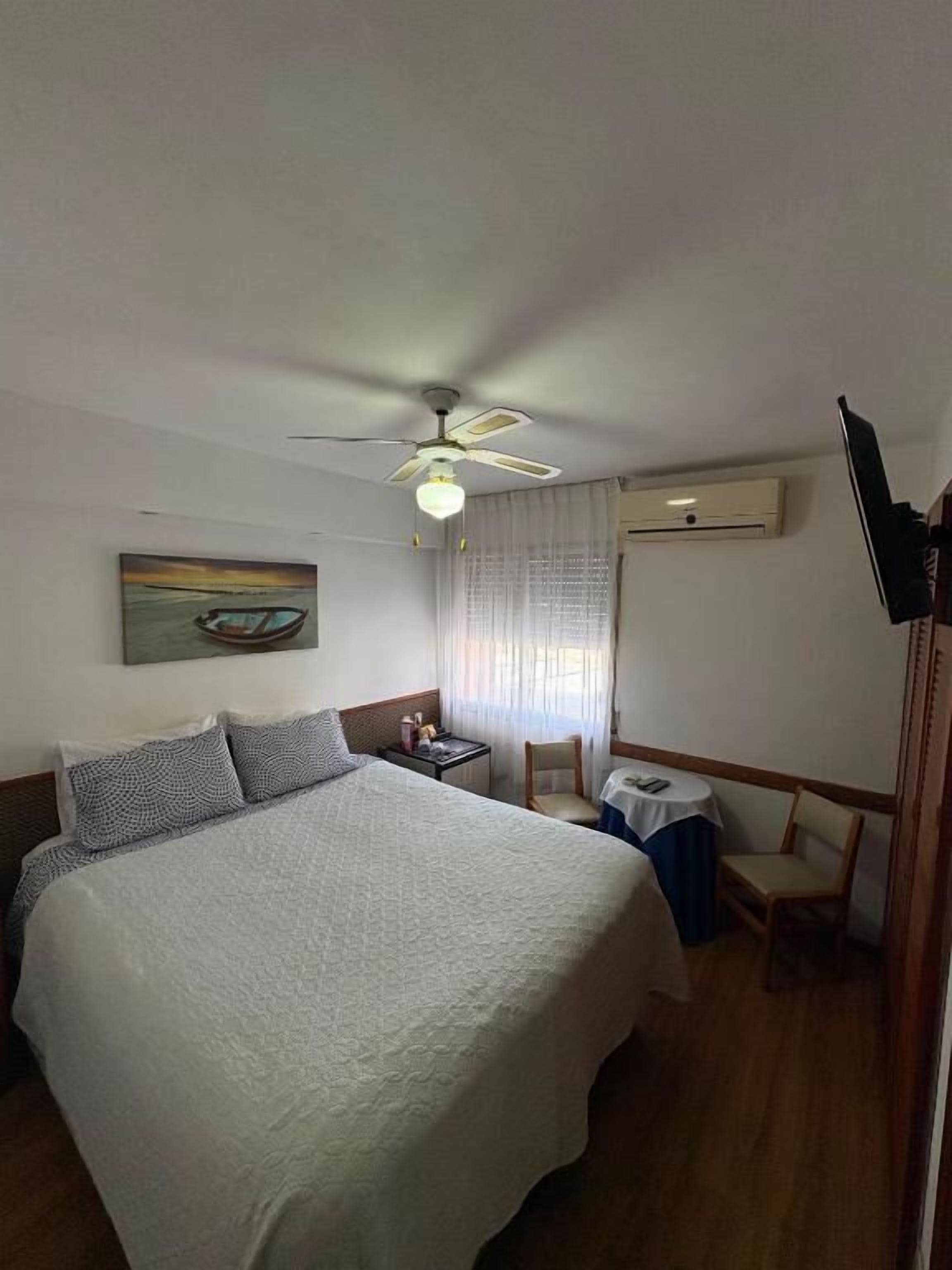 A hotel room with a bed, a desk, a chair, a ceiling fan, and a painting on the wall.