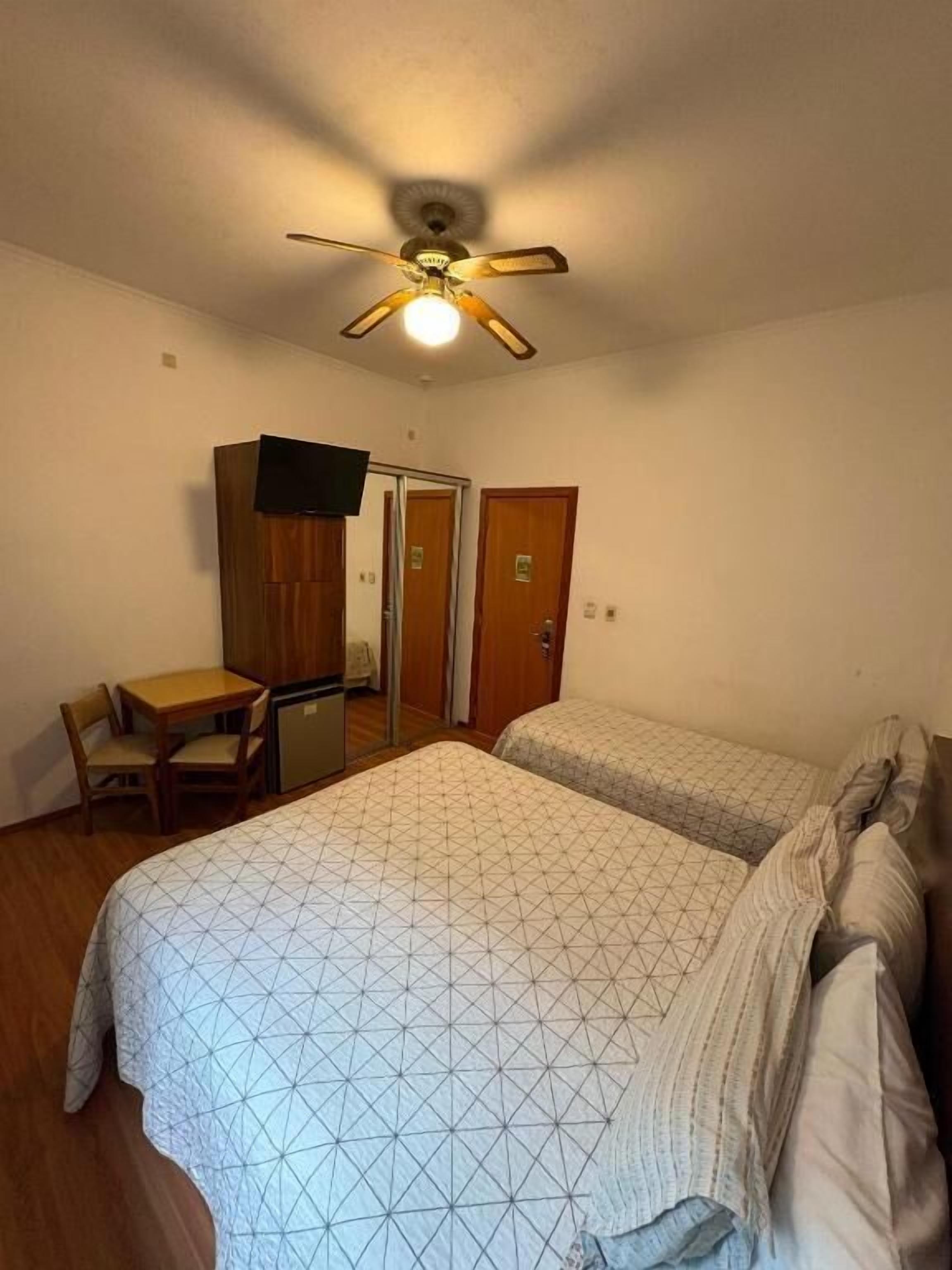 Una stanza con due letti, un ventilatore a soffitto, una scrivania e una sedia.