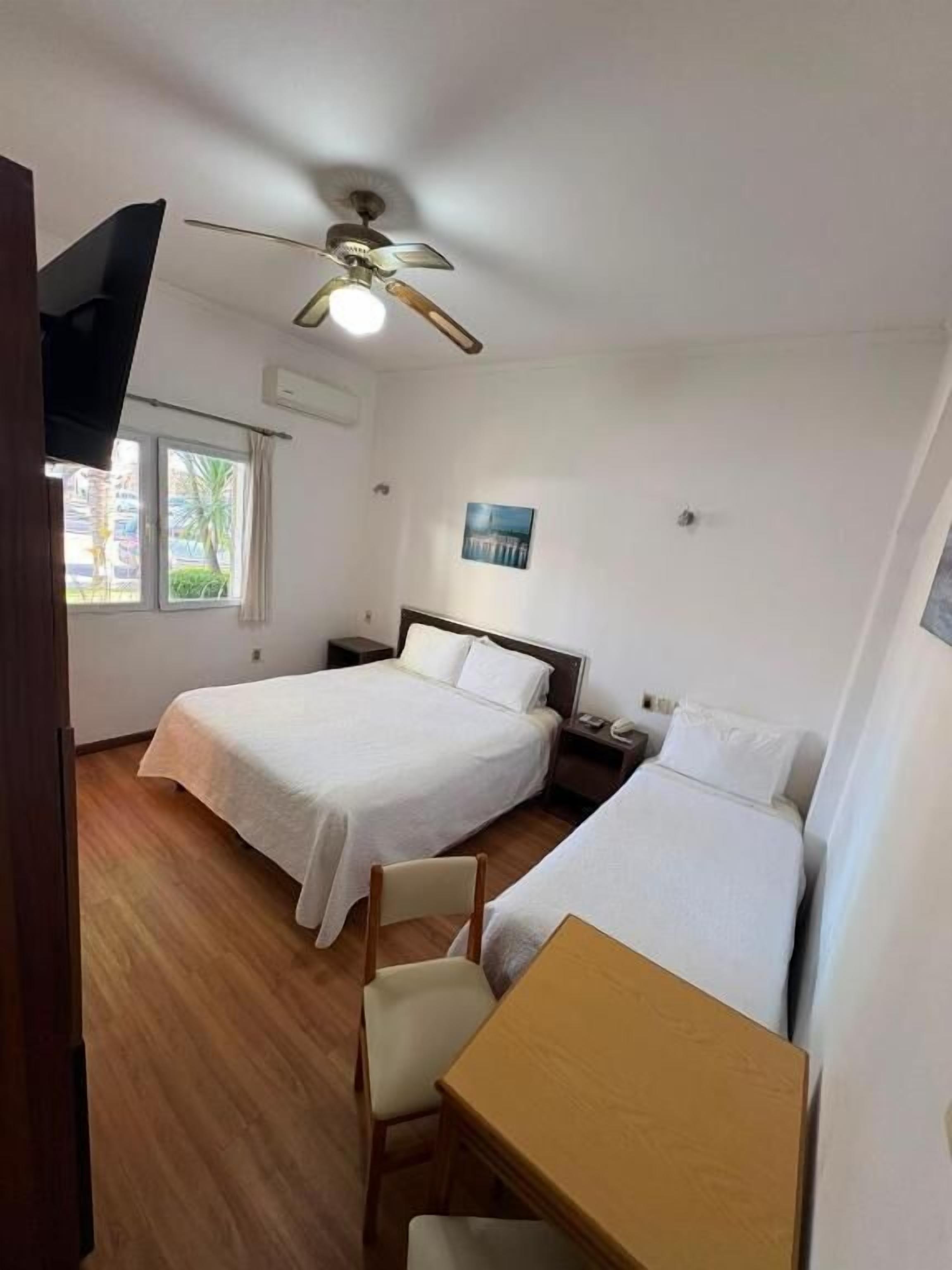 A room with two beds, a wooden table, a ceiling fan, and a window with curtains.