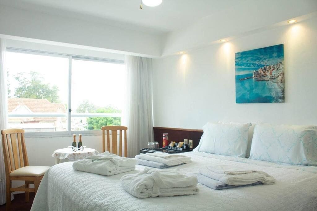 A hotel room with a bed, a chair, a small table with bottles, and a painting on the wall.