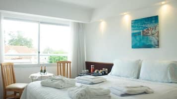A hotel room with a bed, a chair, a small table with bottles, and a painting on the wall.