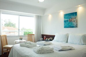 A hotel room with a bed, a chair, a small table with bottles, and a painting on the wall.