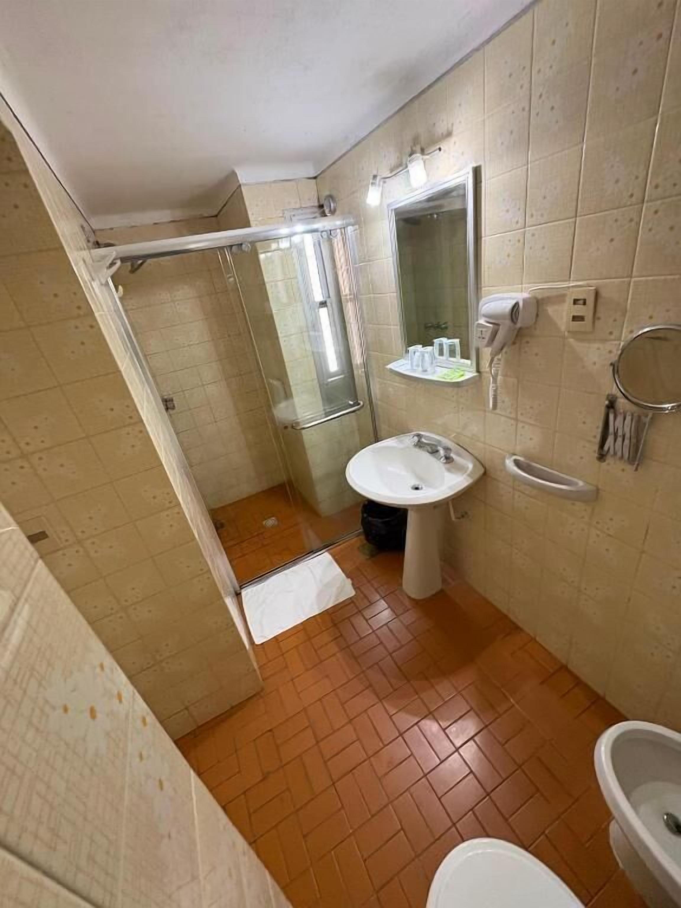Un baño con cabina de ducha de vidrio, lavabo pedestal, espejo y secador de pelo en un estante de pared.