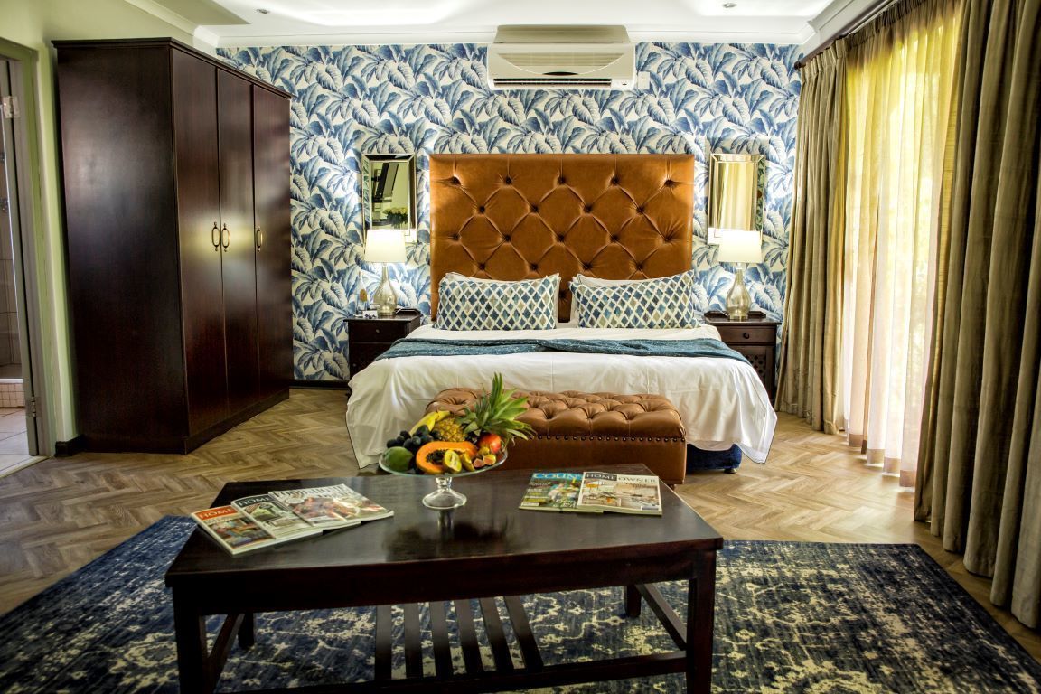 A hotel room with a tufted headboard, a wooden wardrobe, a bed with patterned bedding, a fruit bowl on the bedside table, and a magazine on the table.