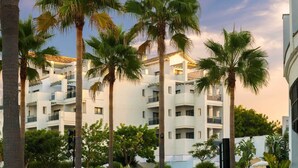 A multi-story building with balconies, surrounded by palm trees and greenery.