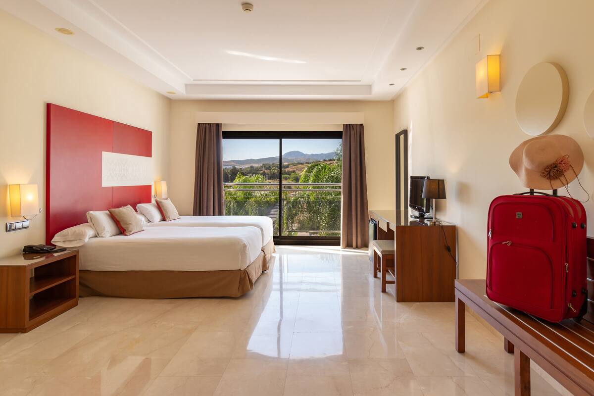 A hotel room with a large bed, a TV, a red suitcase, and a balcony with a view of the hills.