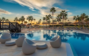 A poolside area with lounge chairs and umbrellas, surrounded by palm trees and a sunset view.