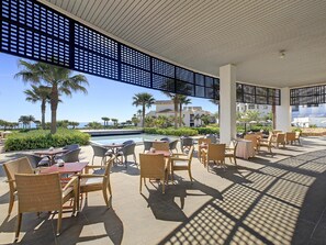 A covered outdoor seating area with tables and chairs, overlooking a pool and palm trees.
