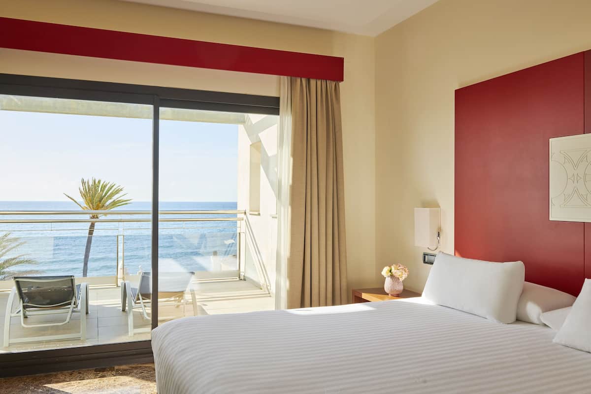 A hotel room with a bed, a nightstand, a lamp, a window with a view of the ocean, and a balcony with chairs and a table.