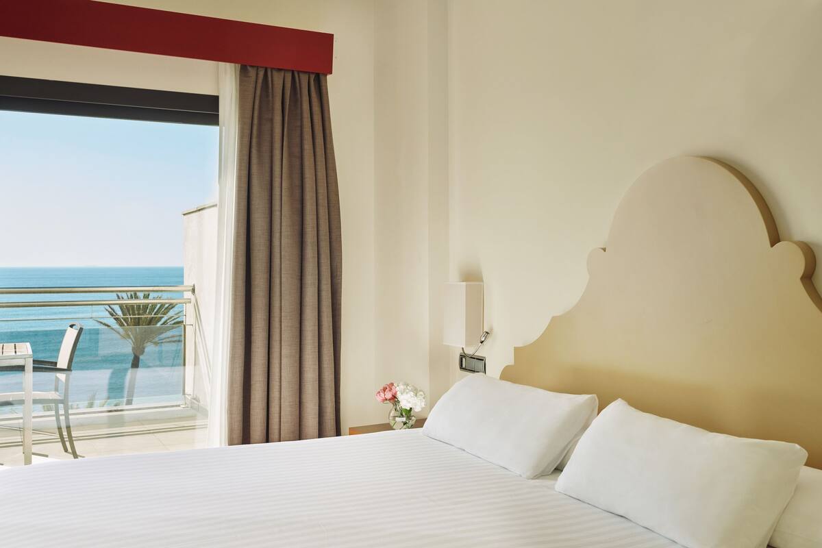 A hotel room with a large bed, a view of the ocean, a balcony with a table and chairs, and a nightstand with a lamp and flowers.