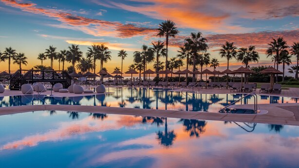 A poolside area with lounge chairs and umbrellas, surrounded by palm trees, reflecting a sunset sky.