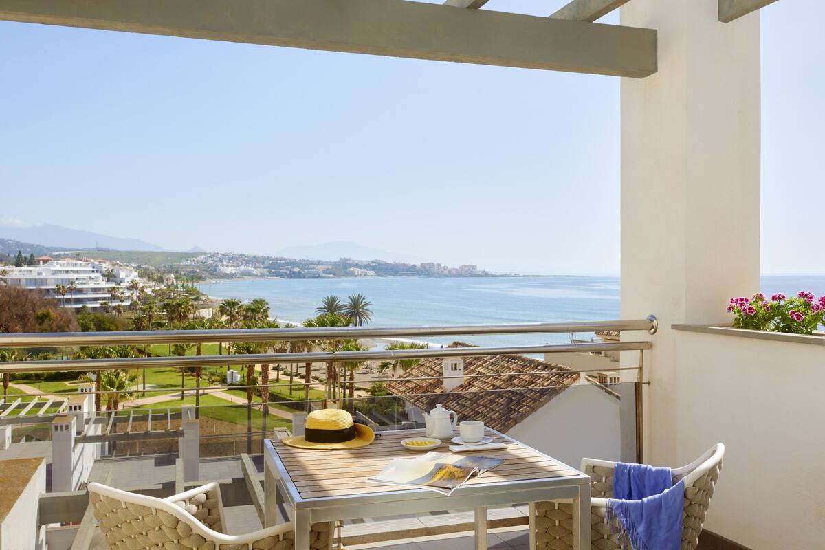 A balcony with a table set for breakfast, overlooking a coastal landscape with buildings and a view of the sea.