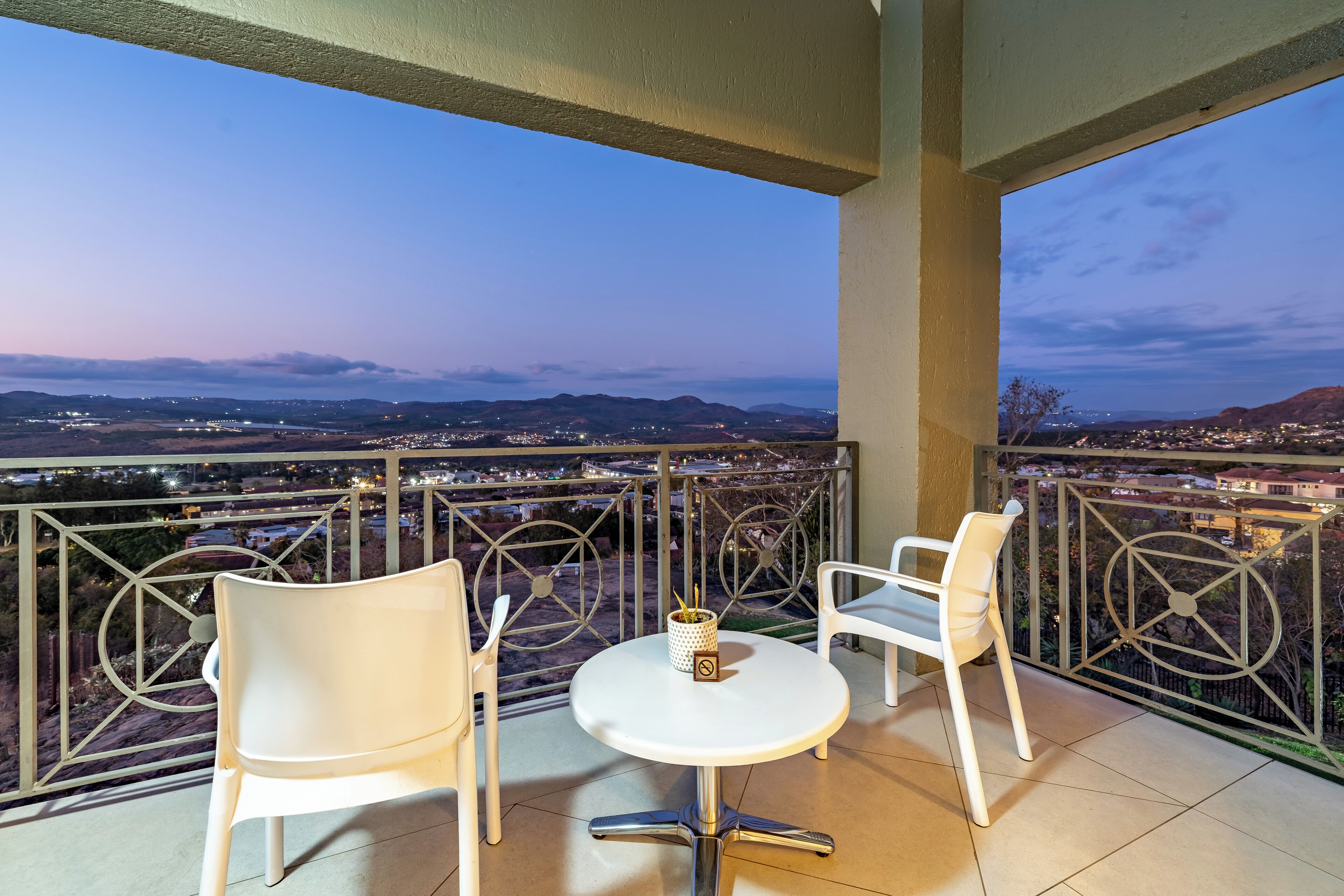 A balcony with two white chairs and a small table, overlooking a cityscape at dusk.