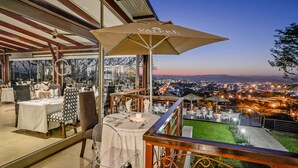 Een terras met tafels en stoelen, een grote parasol en uitzicht op een stadsbeeld in de schemering.