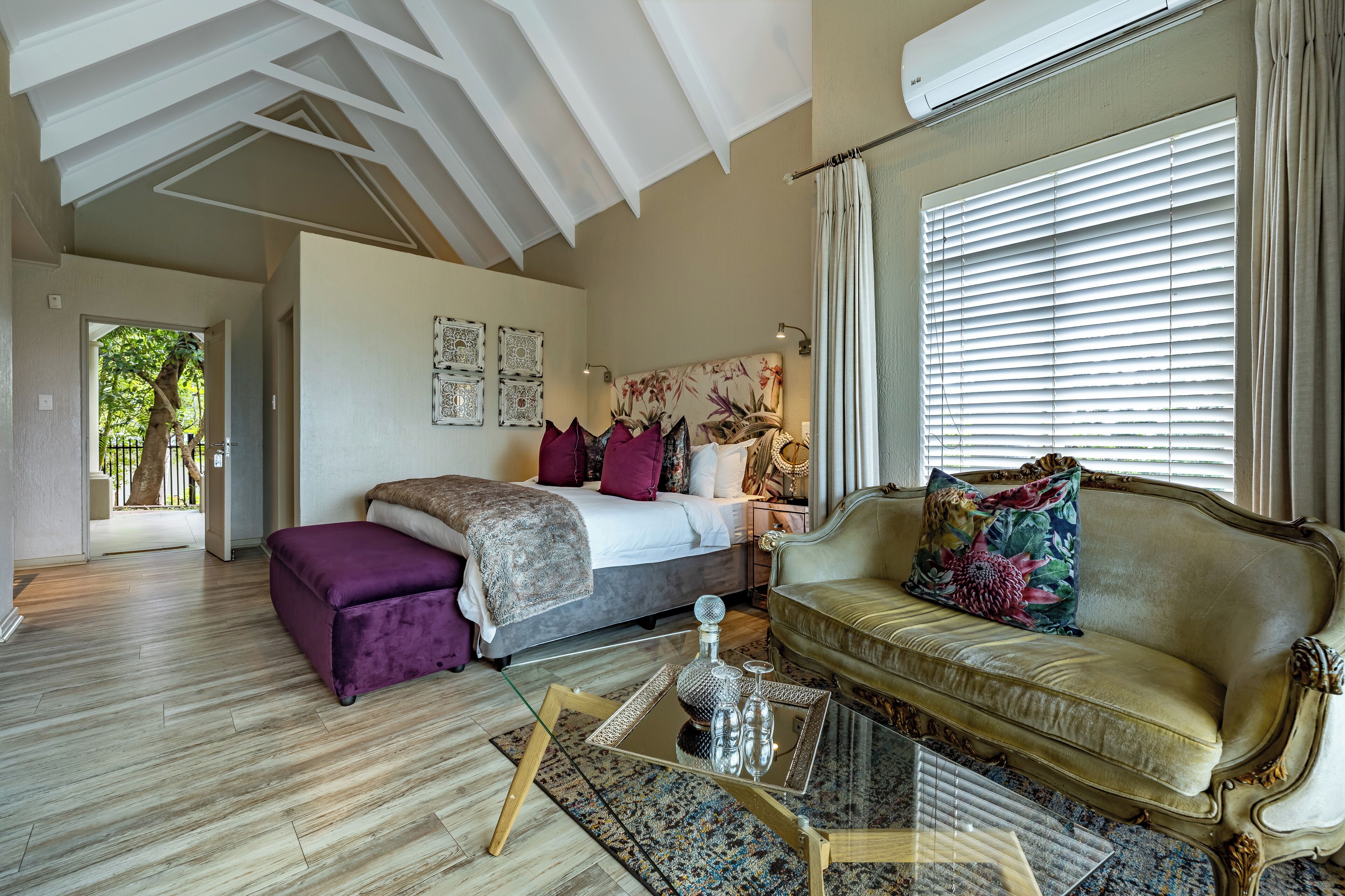 Ruime kamer met een groot bed, een bank en een glazen bijzettafeltje. Er is een raam met zonwering en een plafondventilator.