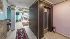 Un vestíbulo de hotel con una mesa de recepción, un extintor, un tapete estampado y una puerta que da a otra habitación.