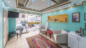 Un vestíbulo de hotel con paredes verde azulado, un techo con diseño y una alfombra roja. Hay dos sillones blancos, una mesita roja pequeña y un televisor montado en la pared.