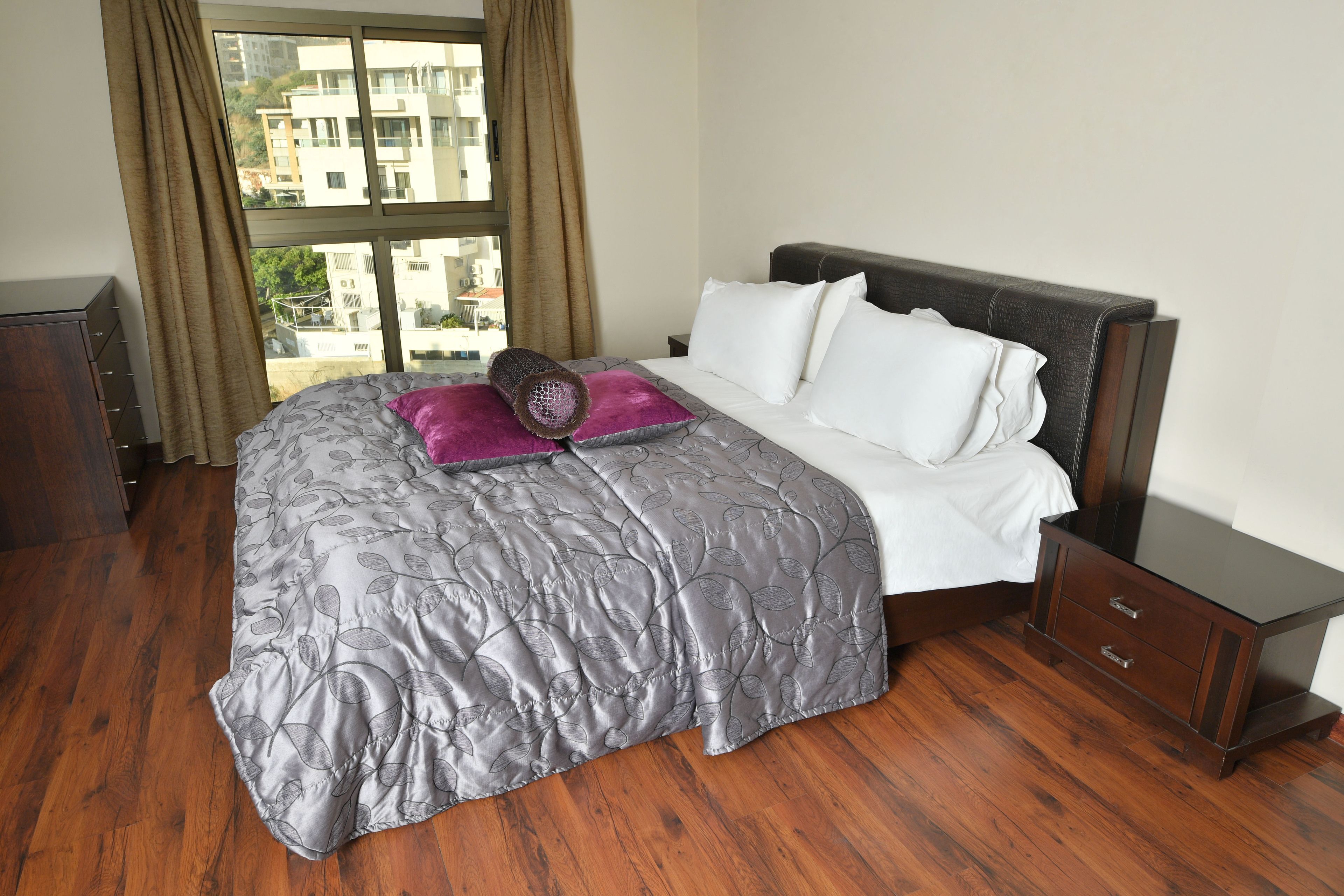 A neatly made bed with a grey floral bedspread and white pillows, a wooden nightstand with drawers, and a window with curtains.