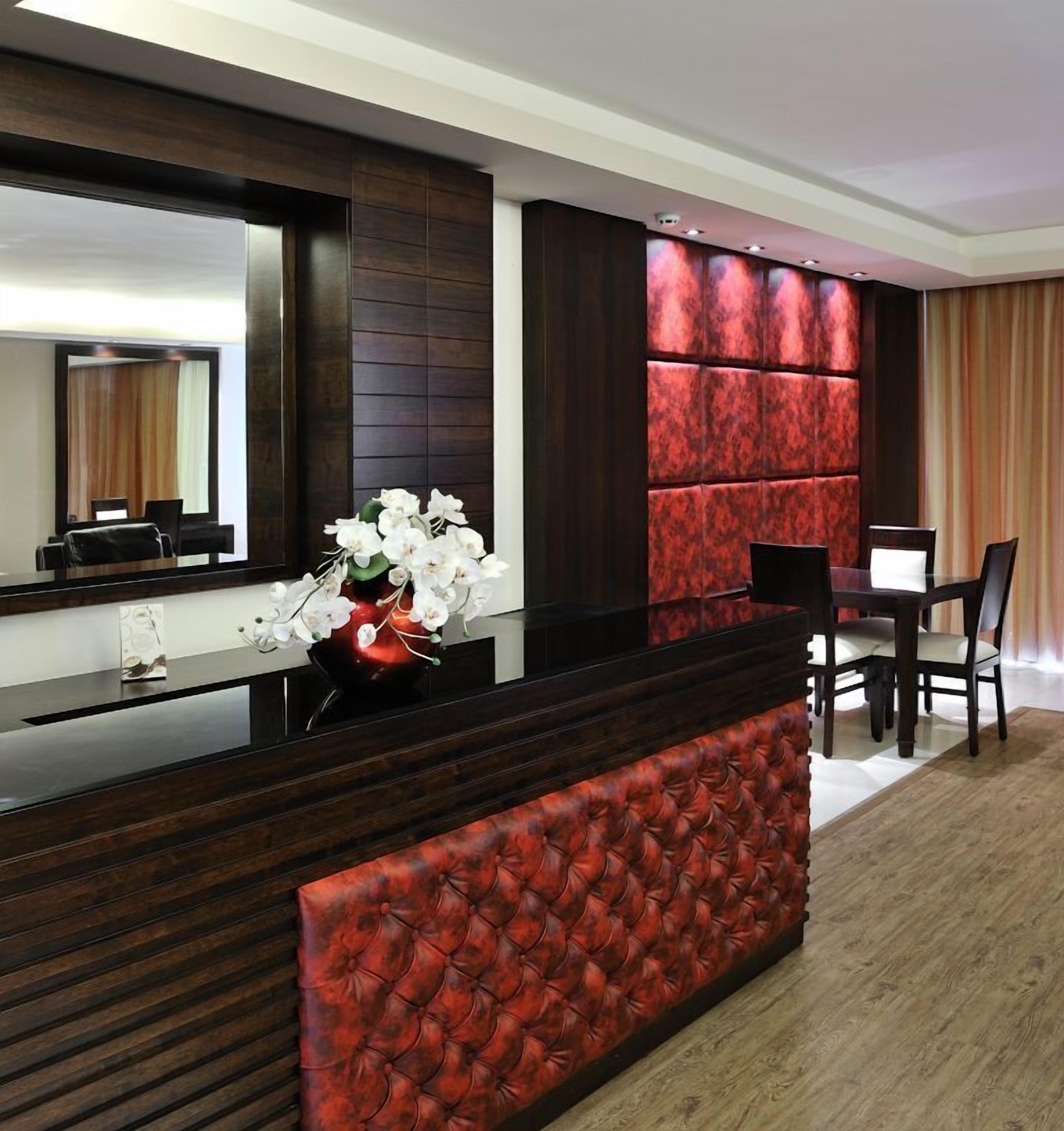 A modern hotel lobby with a dark wood reception desk, a red upholstered bench, and a large mirror.