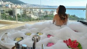A person sitting in a bathtub filled with bubbles and flowers, overlooking a cityscape and the sea. - Hollywood Inn Hotel (Ghazir)