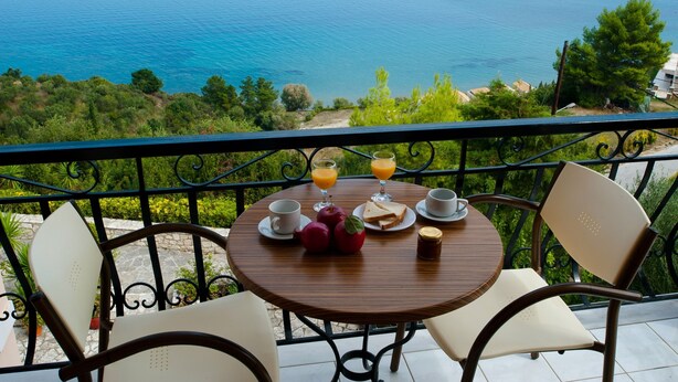 A round wooden table with a breakfast setting on a balcony overlooking a body of water. - Villa Contessa (Zakynthos)