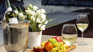 A table with a bottle of wine, a pitcher, a bowl of fruit, and a glass of wine, set against a backdrop of a sunset over water with a ship in the distance. - Artur Hotel (Çanakkale)