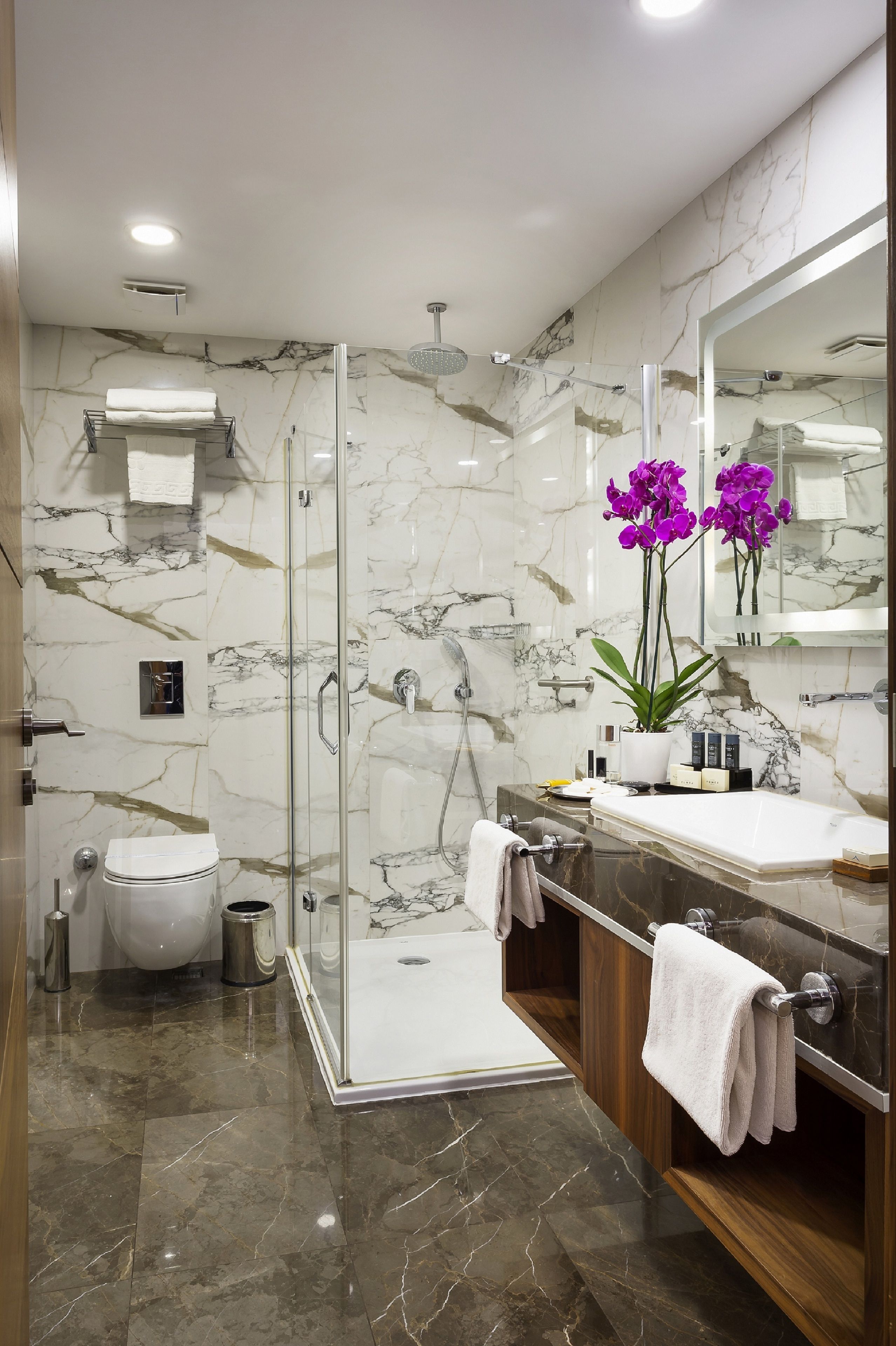 A modern bathroom with a glass-enclosed shower, a marble-tiled floor, a wooden vanity, and a large mirror.