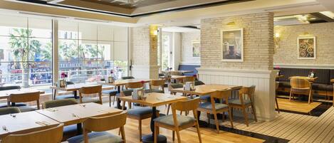 A modern restaurant with wooden tables and chairs, large windows, and a brick accent wall.