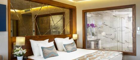 A modern hotel room with a large bed, a wooden headboard, a nightstand with a lamp, and a large window with blinds.