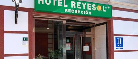The entrance to Hotel Reyes with a reception area, a balcony with a green railing, and a sign indicating the hotel's name and reception.