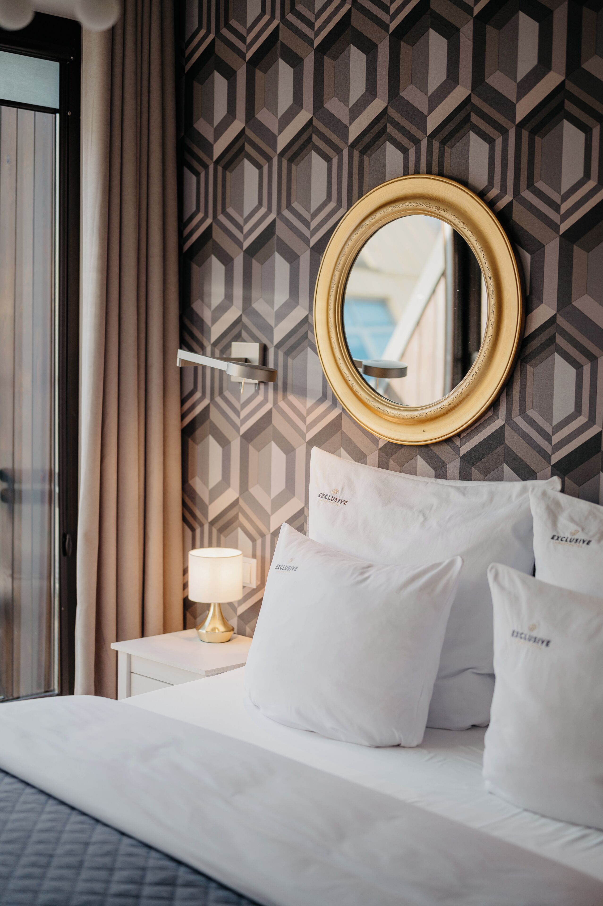 A hotel room with a bed, white pillows, a round mirror, a lamp, and a geometric patterned wall.