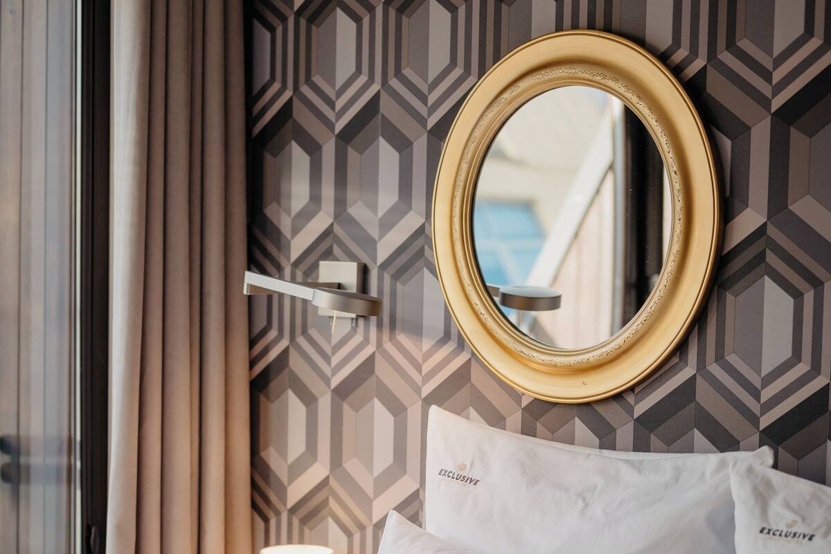 A hotel room with a bed, white pillows, a round mirror, a lamp, and a geometric patterned wall.
