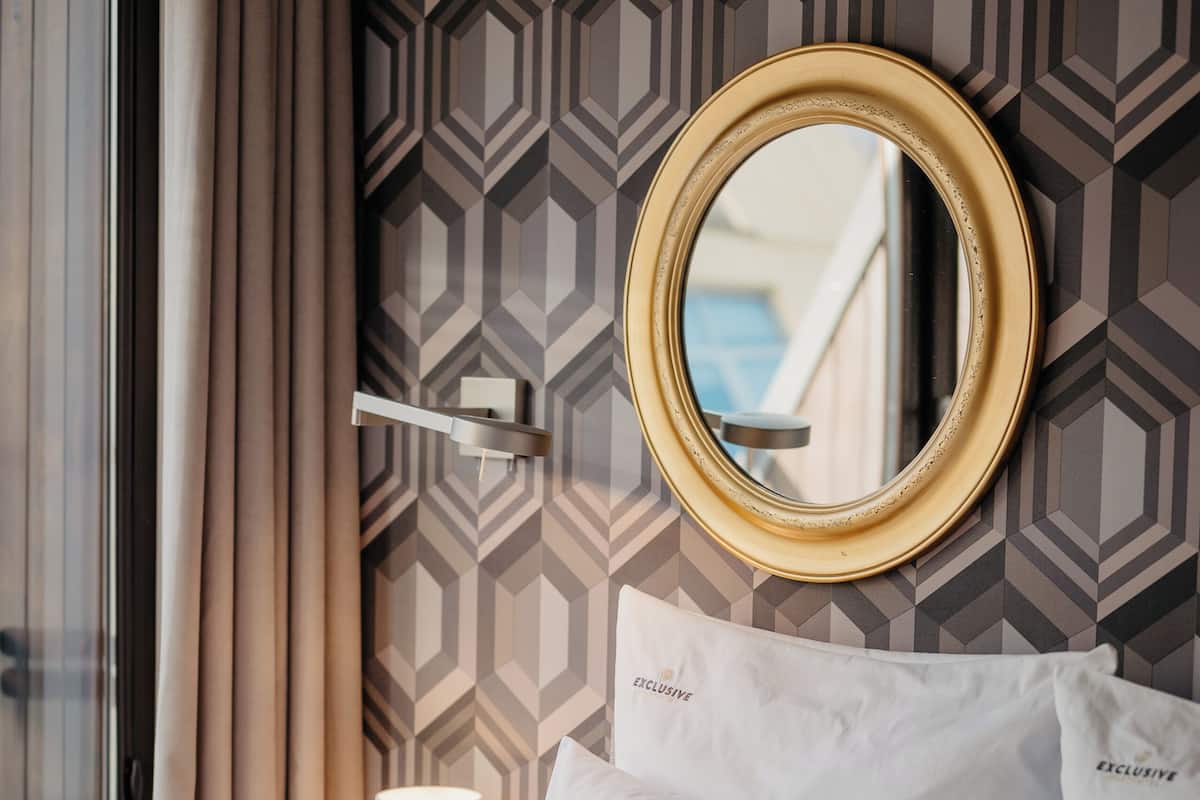 A hotel room with a bed, white pillows, a round mirror, a lamp, and a geometric patterned wall.