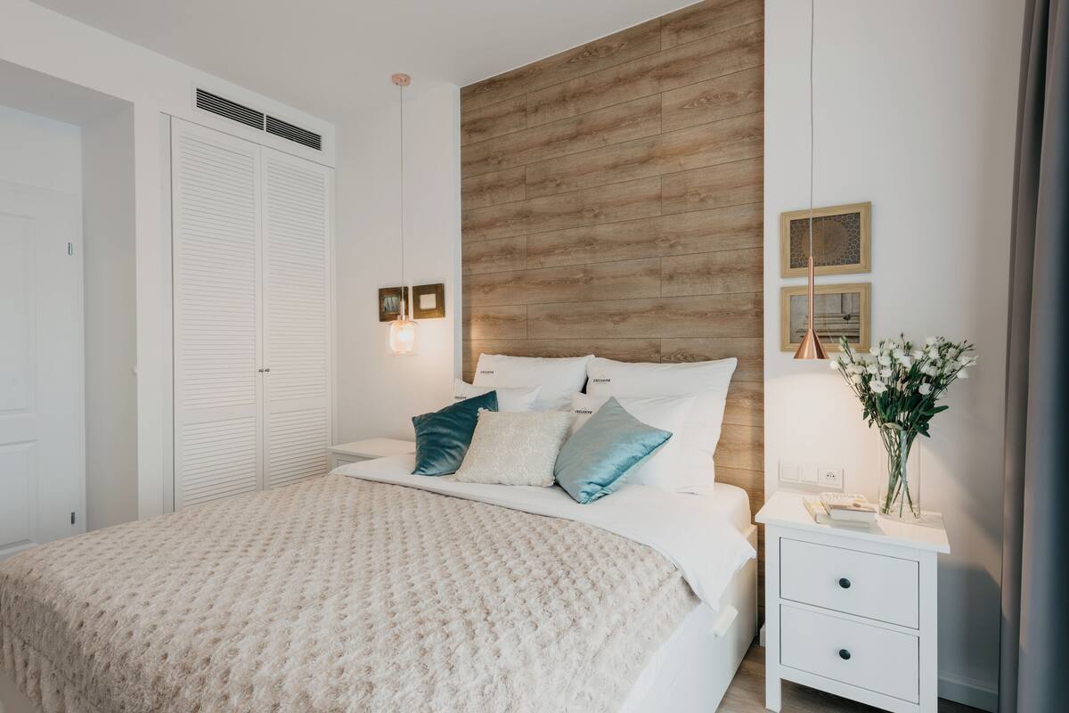 A bedroom with a large bed, white nightstands, a wooden accent wall, and a closet with louvered doors.