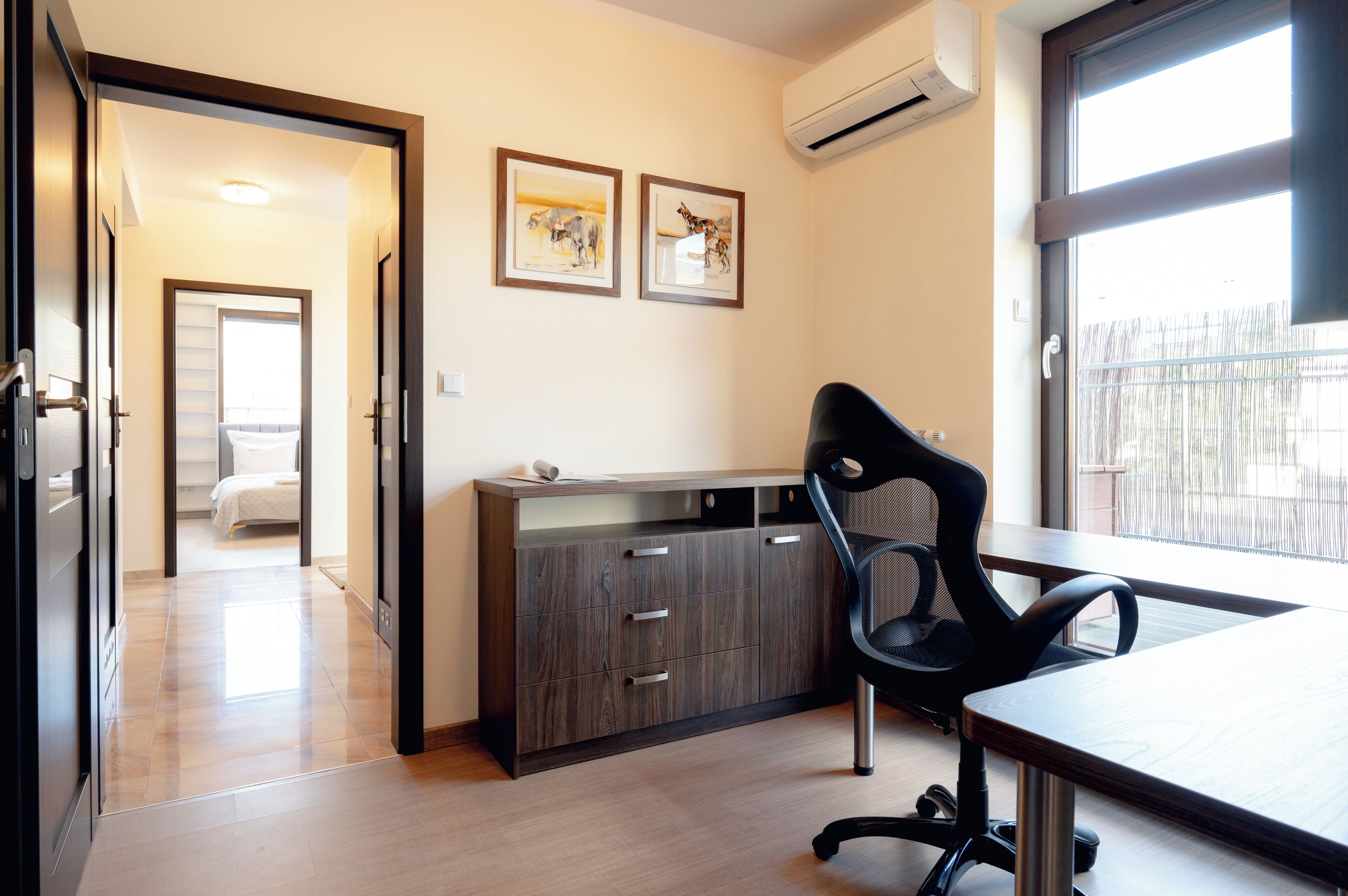 A modern hotel room with a wooden floor, a desk with a chair, a wardrobe, and two framed pictures on the wall.