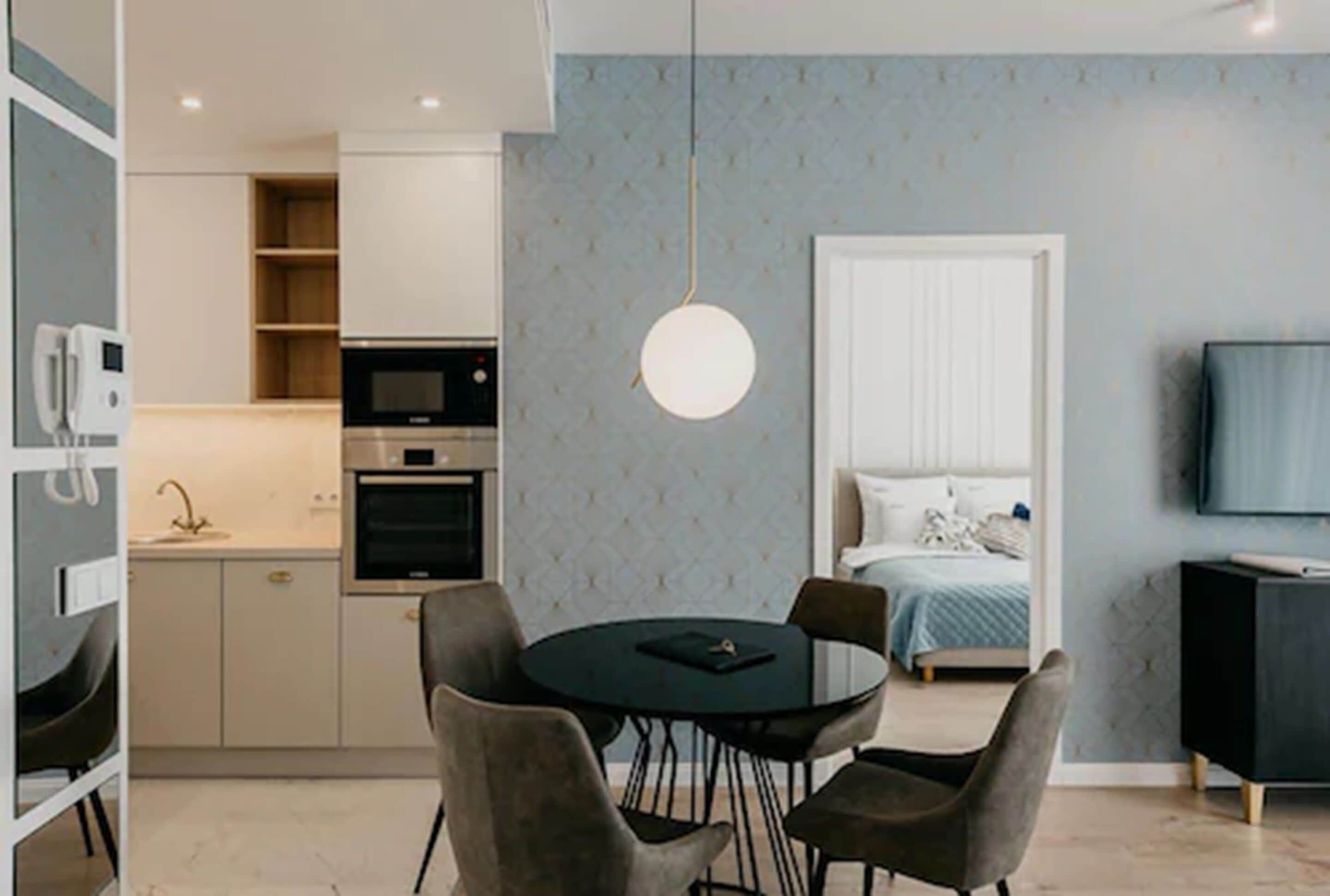 A compact living space with a kitchenette, a dining area with a round table and chairs, a bed with a blue and white duvet, and a television on the right.
