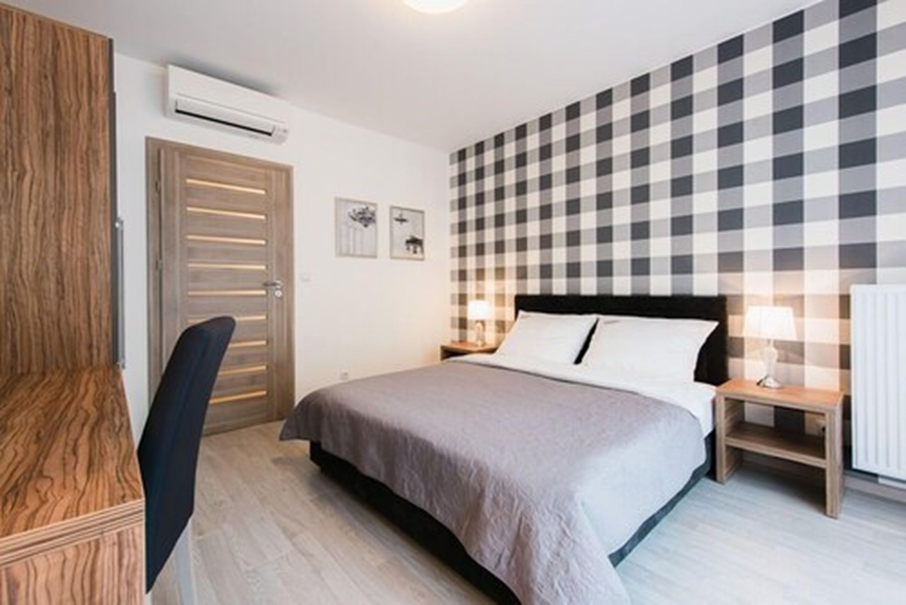 A bedroom with a checkered wall, a bed with a grey bedspread, a wooden wardrobe, a desk, and a chair.