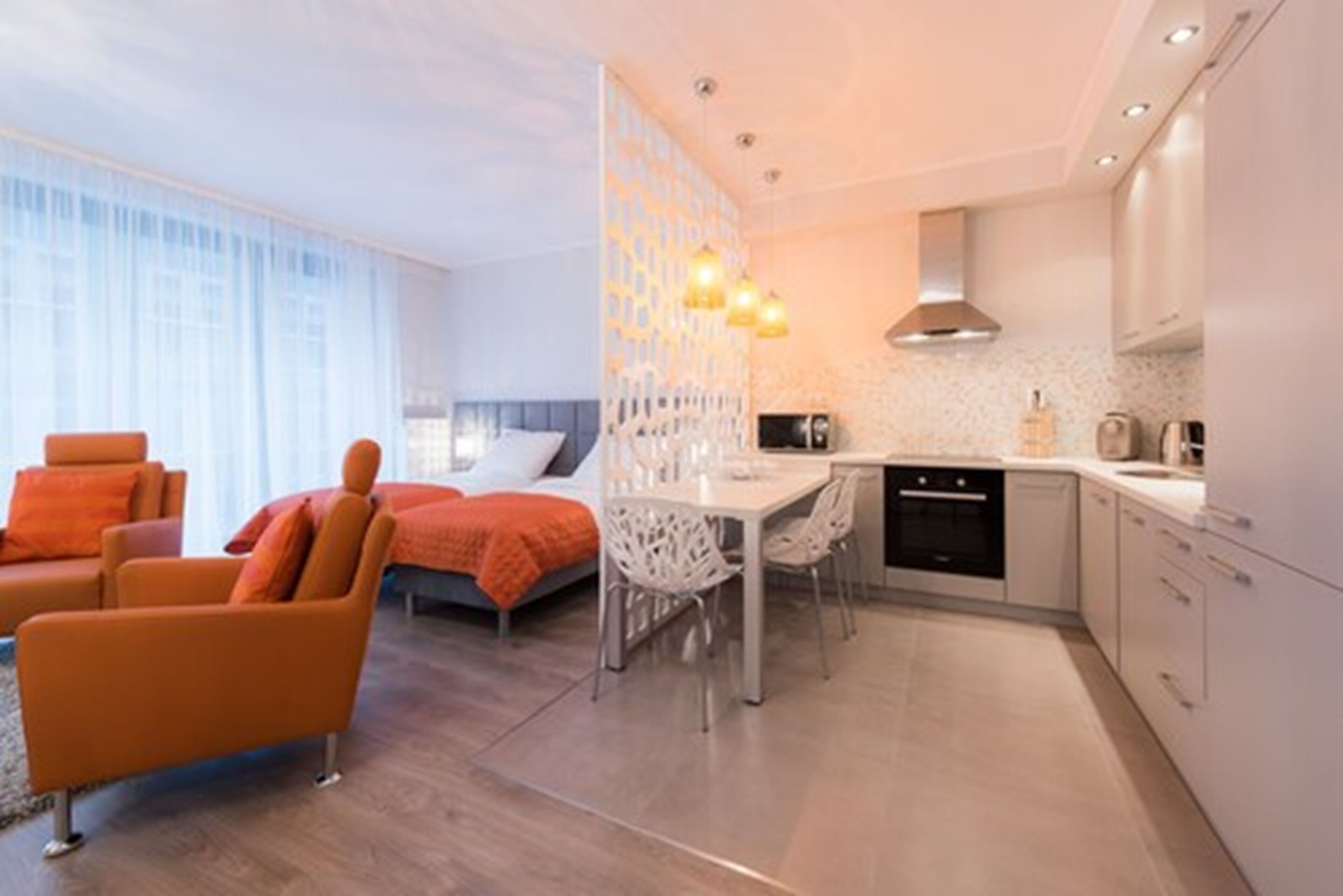 A modern hotel room with a dining area, a bed with orange bedding, and a kitchen with white cabinets.